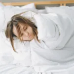 Vrouw slaapt ontspannen in een lichte slaapkamer, helemaal toegedekt onder een dikke witte deken
