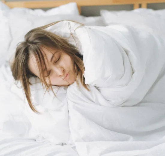 Vrouw slaapt ontspannen in een lichte slaapkamer, helemaal toegedekt onder een dikke witte deken